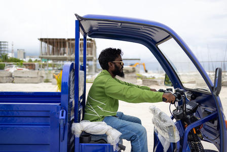 Man in a green shirt driving a blue electric vehicle by the harbor.