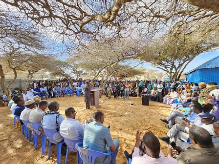 Outdoor community gathering under trees with blue chairs around a central podium.