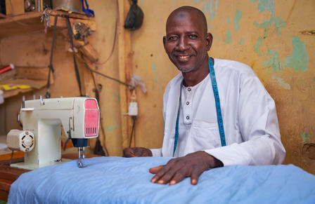 Photograph of a tailor at a sewing table with blue fabric in a rustic workshop