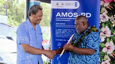Two men shake hands outdoors beside an AMOS-RD banner and a flower backdrop.