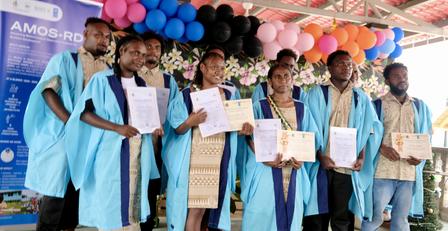 Photograph of graduates in blue gowns holding certificates on stage with balloons.