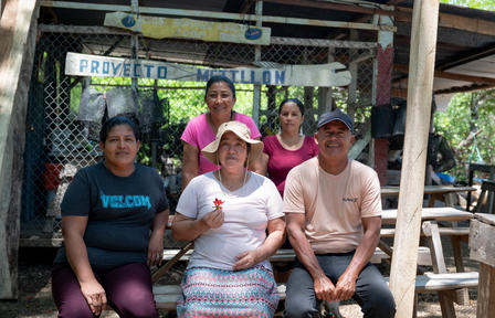 Five people sitting on a wooden bench under a shelter, smiling at the camera.