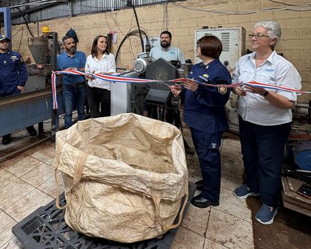 People in a workshop cut a red-and-white ribbon during a ceremony; brown paper bag on table.