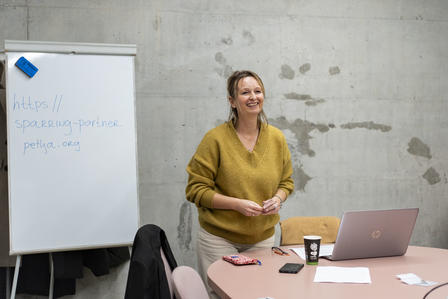 Woman in mustard sweater stands by a whiteboard in a gray room; pink table with laptop.