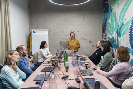 Photograph: Group of coworkers around a conference table as a woman presents at the front.