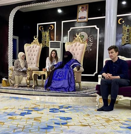 People sit on ornate gold chairs on a luxury TV set with purple decor.