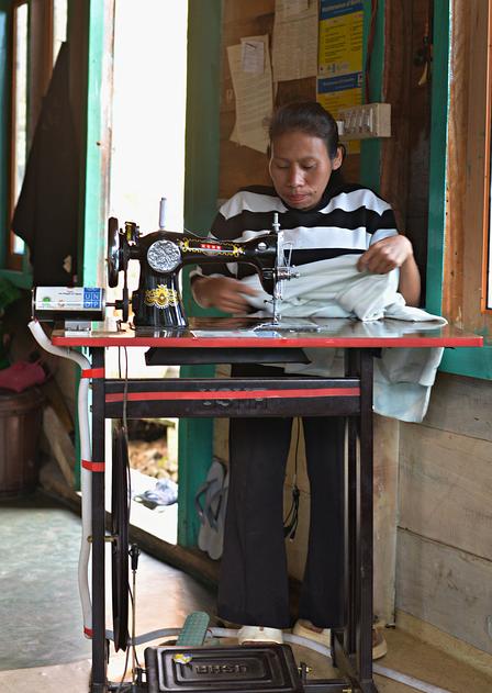 Seamstress sits at a sewing machine on a red table inside a small workshop.