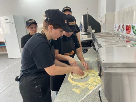 Team of cooks in black uniforms roll out dough on a stainless prep table in a busy kitchen.