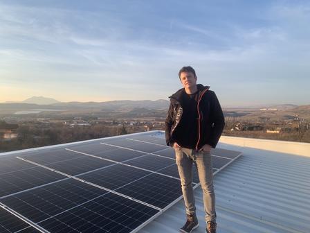 Young man standing on a metal rooftop with solar panels, overlooking a valley at sunset.