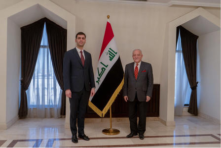 Two men in dark suits stand on a marble floor beside the Iraqi flag in a formal room.