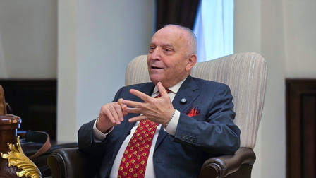 Man in a dark blue suit with red tie sits in an upholstered chair, gesturing with his hands.