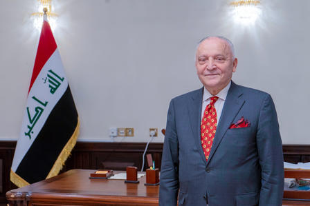Man in blue suit and red tie stands in a formal room beside the Iraqi flag.
