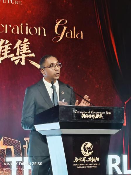 Man in a suit speaks at a podium on stage with a red gala backdrop.