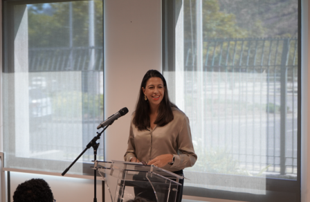 Photograph of a woman speaking at a podium with a microphone in a bright room.