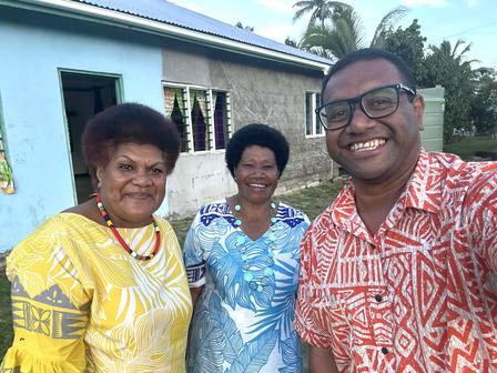 Three people in bright patterned shirts pose in front of a blue building outdoors.