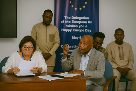Two men in suits signing documents at a table; a standing man behind them; blue banner behind.