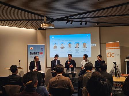 Panel discussion on stage with several speakers; audience seated, screen shows headshots.