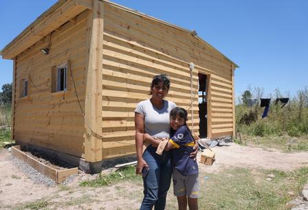 Two people smile in front of a small wooden log cabin under construction on a sunny day.