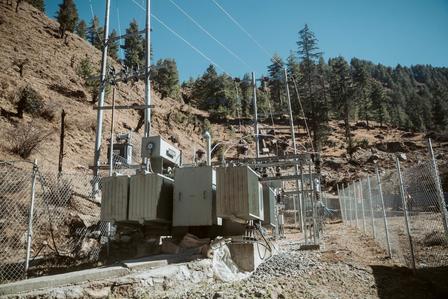Utility transformers and electrical equipment behind a chain-link fence on a hillside with trees.