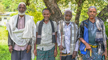 Four men in traditional attire stand together outdoors among trees, smiling.