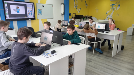 Several students sit at white desks, using laptops; bright room with a yellow wall.