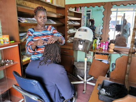 Hairstylist stands behind a seated client in a small salon, with shelves of hair products.