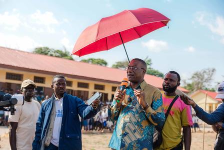 Group of men outdoors; man in blue patterned shirt speaks into microphone under a red umbrella near a yellow building.