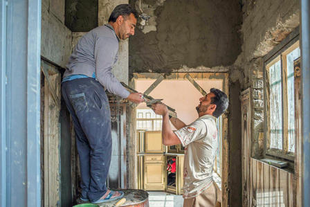 Two men renovate a dilapidated room, passing a wooden beam between them.