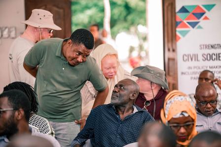 Group at an outdoor event; man in a green shirt leans toward a seated man near a Civil Society banner.