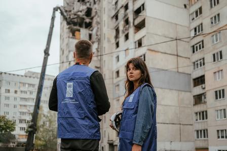 Two people in denim jackets stand near a partially burned high-rise in an urban neighborhood.
