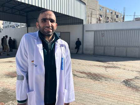 Photo of a person in a white lab coat and green scarf standing outdoors near a brick building.