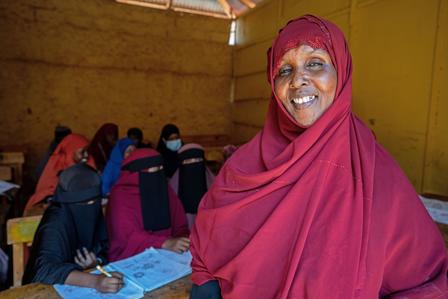 Women in colorful hijabs sit at desks in a yellow-walled classroom; foreground pink hijab.