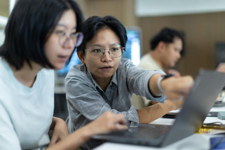 Three students in a classroom working on laptops at a table.