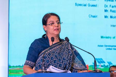 Speaker in a blue patterned outfit at a podium with a microphone, blue screen behind.