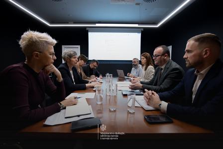 Photo: business meeting around a long conference table with papers and water bottles; screen.