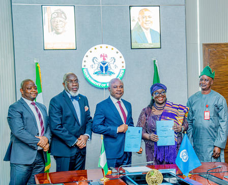 Photo of six people in formal attire at a signing ceremony with Nigerian flags in the background.