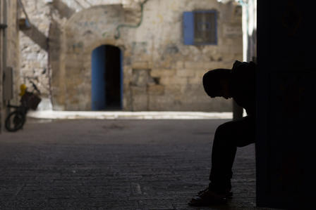 Silhouette of a person sitting in a dark doorway on a sunlit stone street, blue arched door.