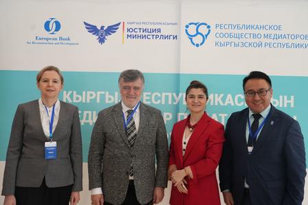 Photograph: four professionals in business attire pose in front of a teal conference banner with logos.
