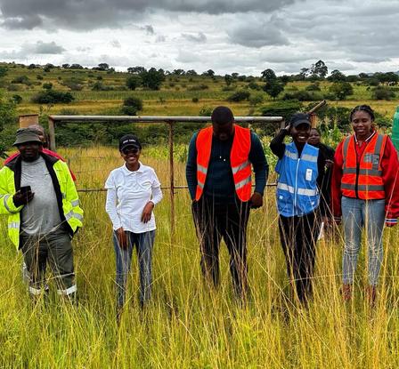 Group of people in reflective vests standing in tall grass by a river in a rural field.