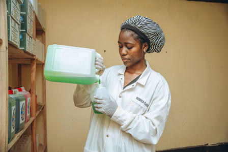 Lab worker in white coat and hairnet holds a green and white container; shelves with bottles.