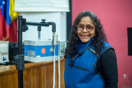 Person in a blue vest beside a blue ballot drop box at a polling station.