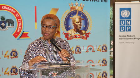 Speaker at podium wearing a checkered shirt on stage with banners and logos in the background.