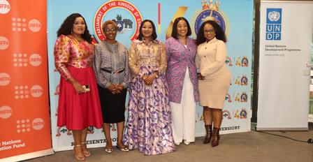Photograph: five women in colorful dresses posing in front of event banners with logos.