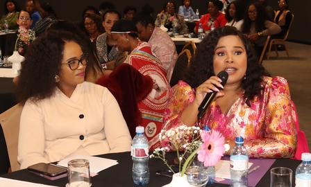 Two women sit at a banquet table; one in a beige cardigan, the other in a pink floral dress.