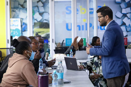 A man in a blue blazer talks to a seated group of people in a training.