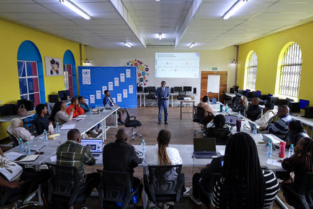 Workshop in a yellow room; participants sit around tables as a presenter speaks near a whiteboard.