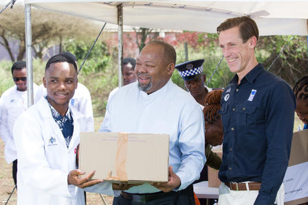 Group of people under a canopy; man in white hands a cardboard box to another man.