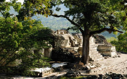 Ancient terraced stone ruins amid trees beside a rocky stream and distant hills.