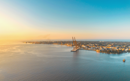 A photograph of a harbor at sunset with cranes and a distant city skyline across calm water.