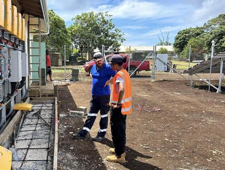 Two construction workers pose with arms around each other on a dirt site, wearing safety gear.